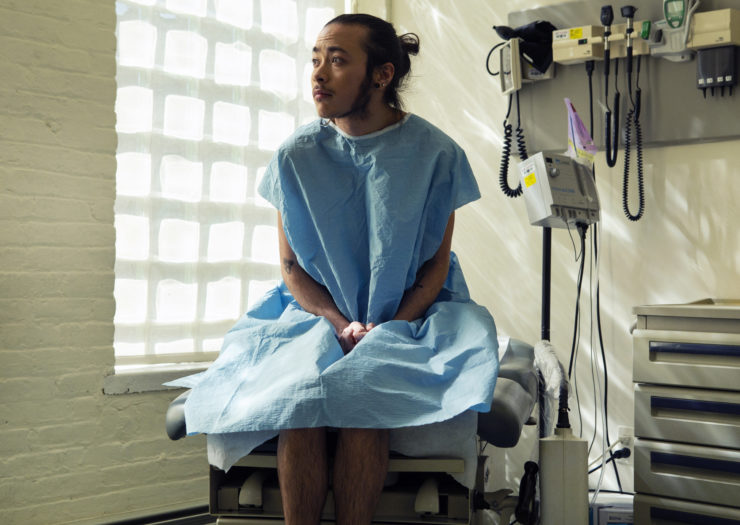 Photo of a a genderqueer person sitting in a hospital gown sitting in an exam room