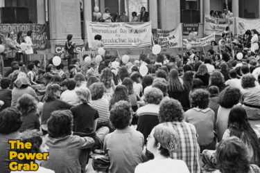 Black and white photo of a large crowd sitting and facing a sign that reads International Day of Abortion