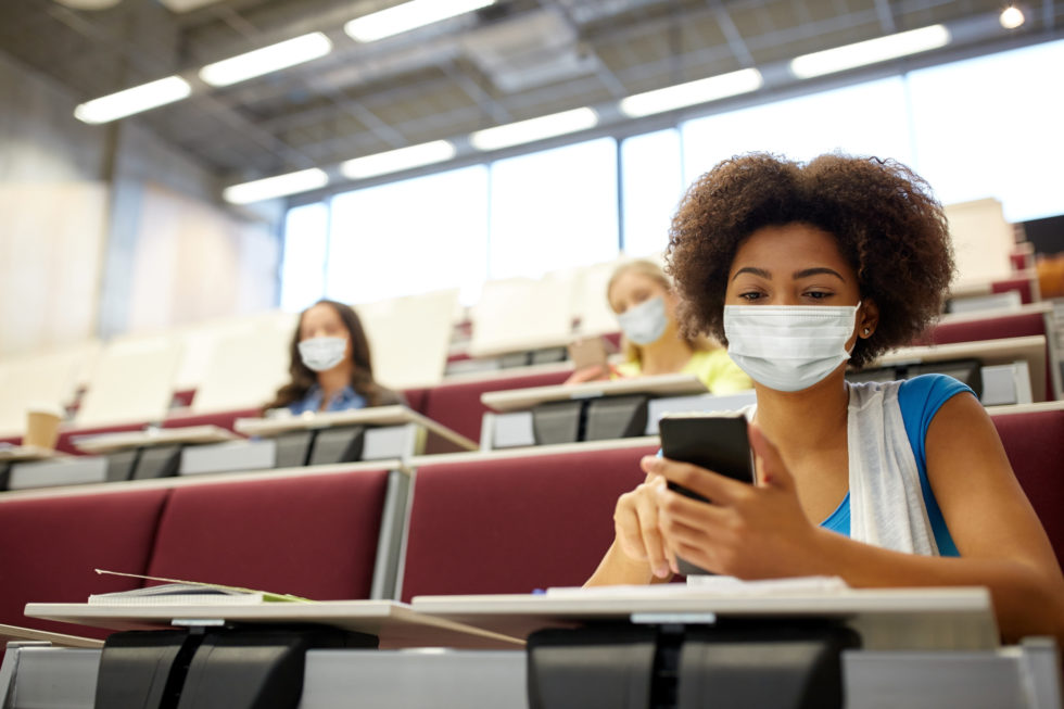 Photo of Black student wearing a face mask sitting in classroom on her phone
