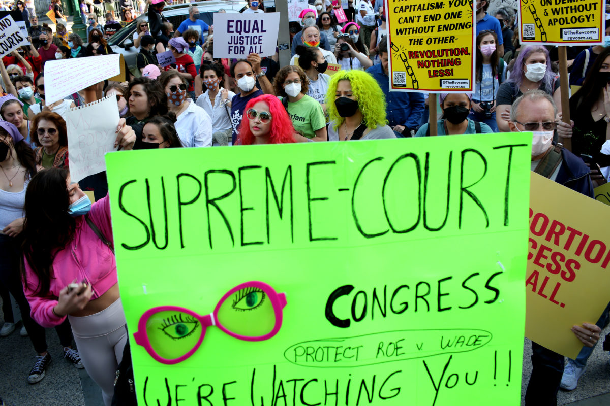 Photo of a crowd of protesters with a neon green sign in front that reads Supreme Court + Congress we're watching you