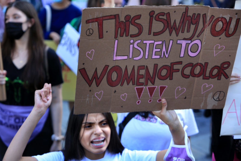 Photo of an abortion rights protester holding a cardboard sign that reads This is why you listen to women of color