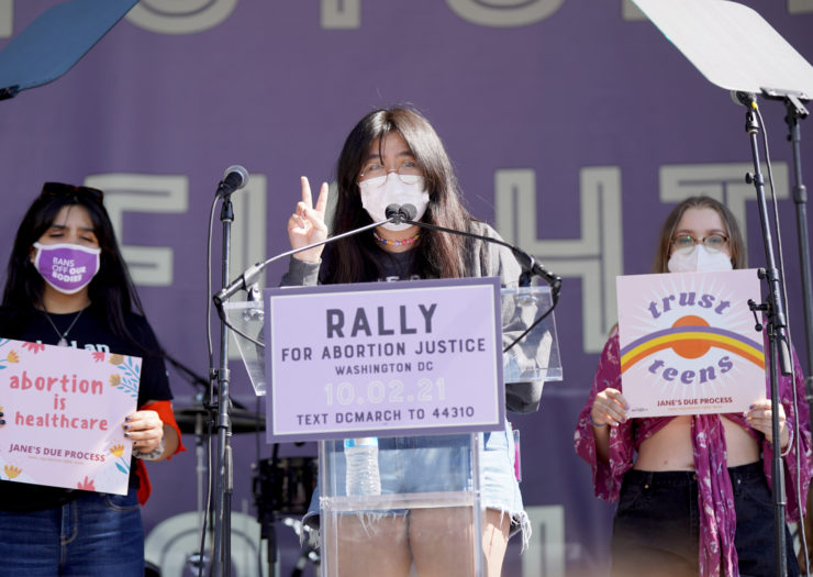 Photo of three teens on stage, one speaking, at the Rally for Abortion Justice