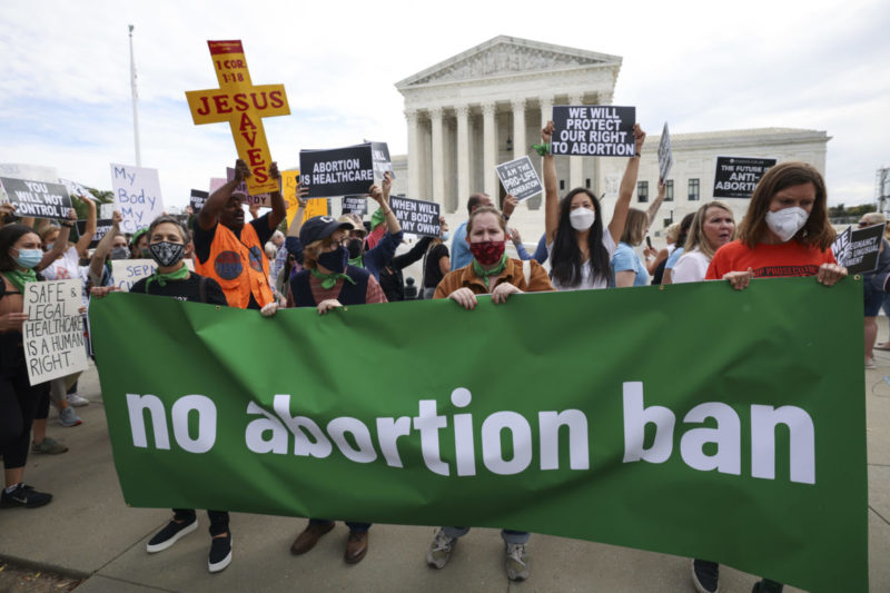 Photo of abortion rights activists outside of the Supreme Court holding a large green sign that reads no abortion ban in white lettering