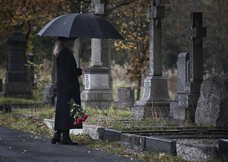 Photo of woman at a grave site in dark clothing, holding roses in one hand and an umbrella in the other