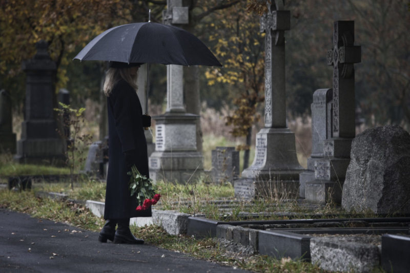 Photo of woman at a grave site in dark clothing, holding roses in one hand and an umbrella in the other