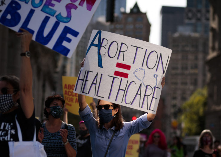 Photo of woman at a protest wearing sunglasses and a mask holding up a sign that says abortion = health care