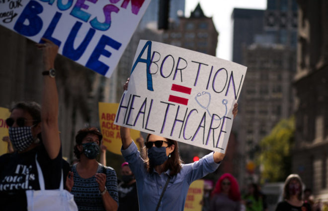 Photo of woman at a protest wearing sunglasses and a mask holding up a sign that says abortion = health care