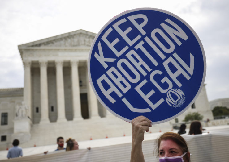 Photo of an abortion rights protester with the Supreme Court in the background holding a blue around sign that says in white lettering keep abortion legal