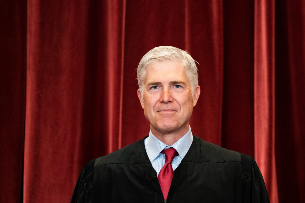 Photo of Associate Justice Neil Gorsuch wearing a black robe, blue collared shirt and red tie in front of a red curtain