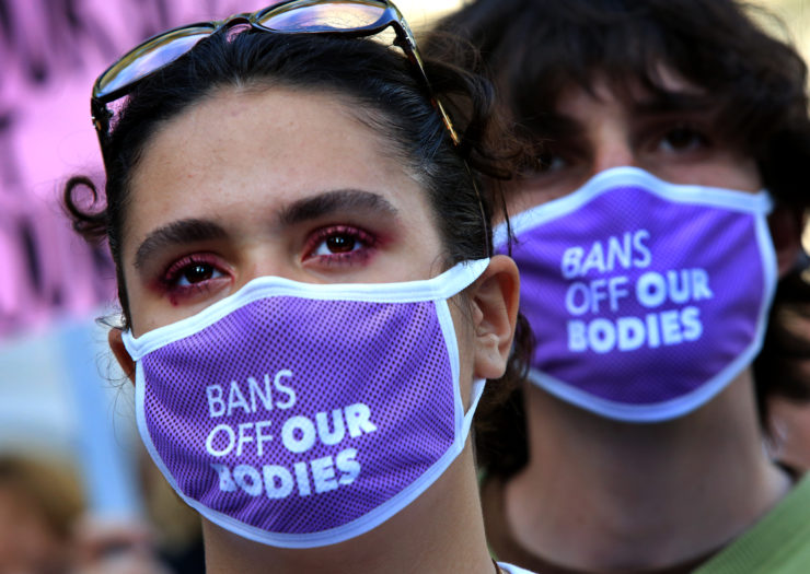 Photo of a closeup of two teens wearing purple masks that say bans off our bodies