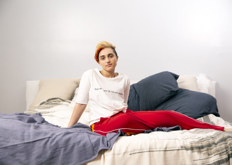 Photo of a transmasculine gender nonconforming person sitting on a bed with blankets and pillows around them