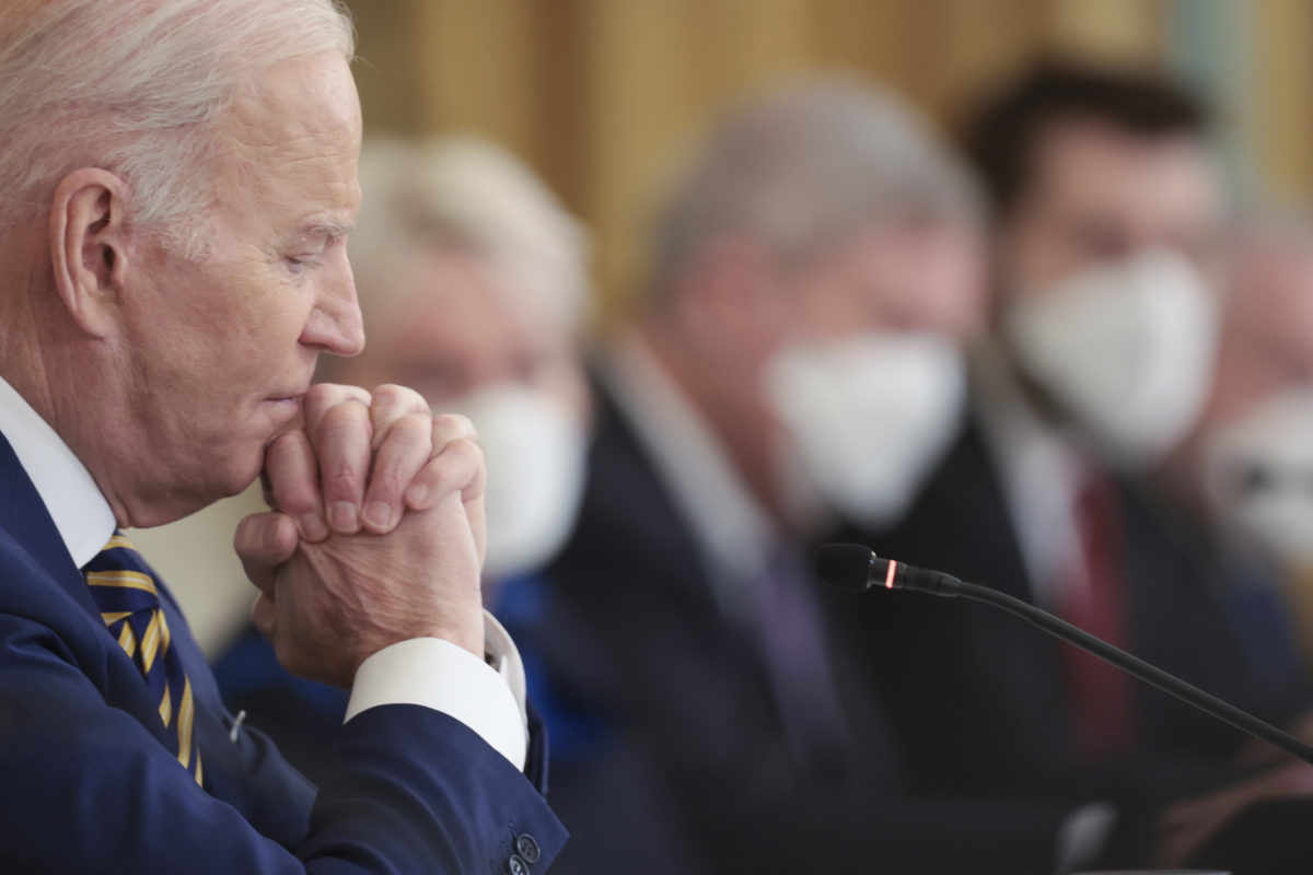 Photo of President Biden in the foreground sitting with his hands folded