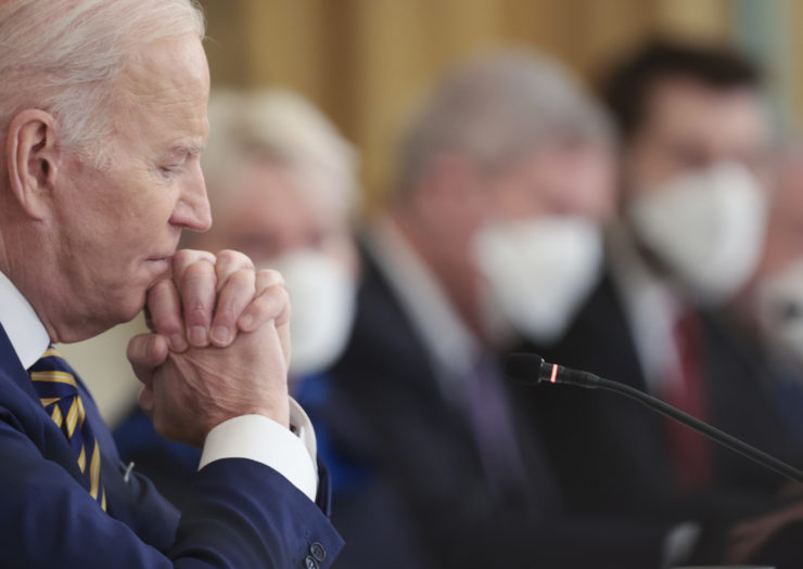 Photo of President Biden in the foreground sitting with his hands folded