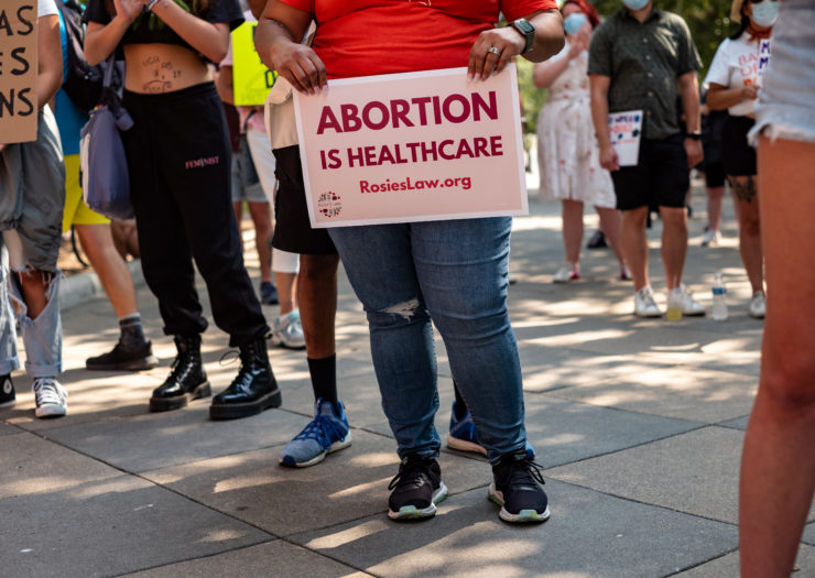 Protest photo of an activist holding a sign that reads Abortion Is Health Care RosiesLaw.Org