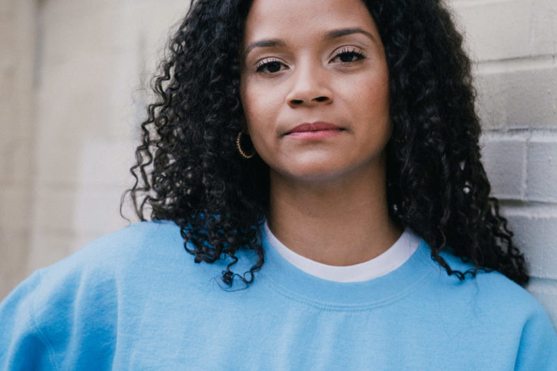 headshot image of Aisha Becker-Burrowes wearing a light blue FEMINIST sweatshirt