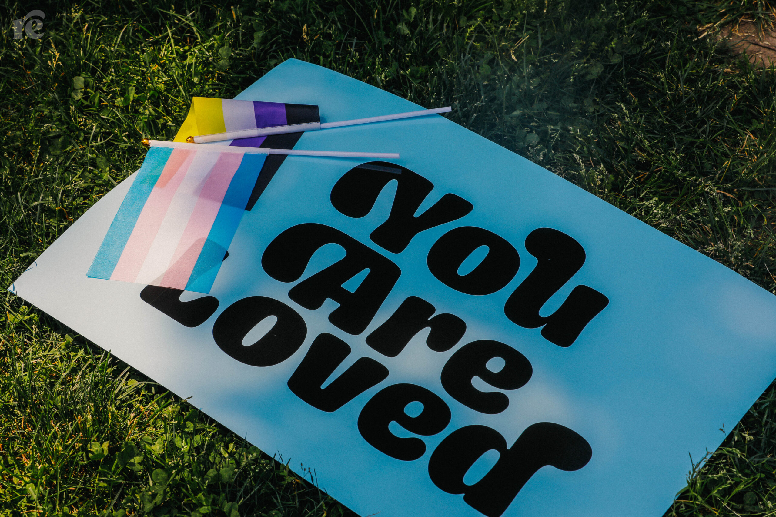 Blue poster with the text "You are loved" and the transgender pride flag and nonbinary pride flag laying on top.