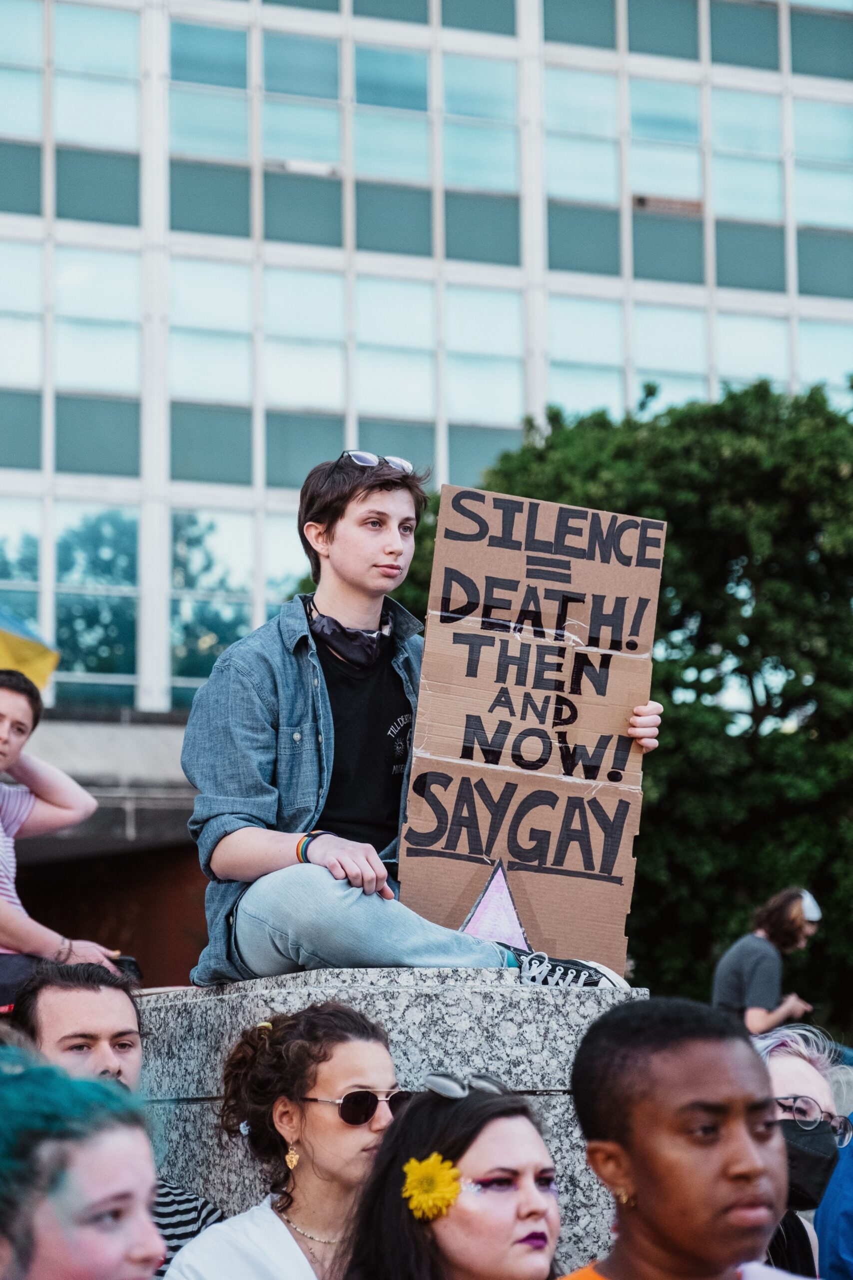 A person holding a sign with the text "Silence is death! Then and now! Say gay."