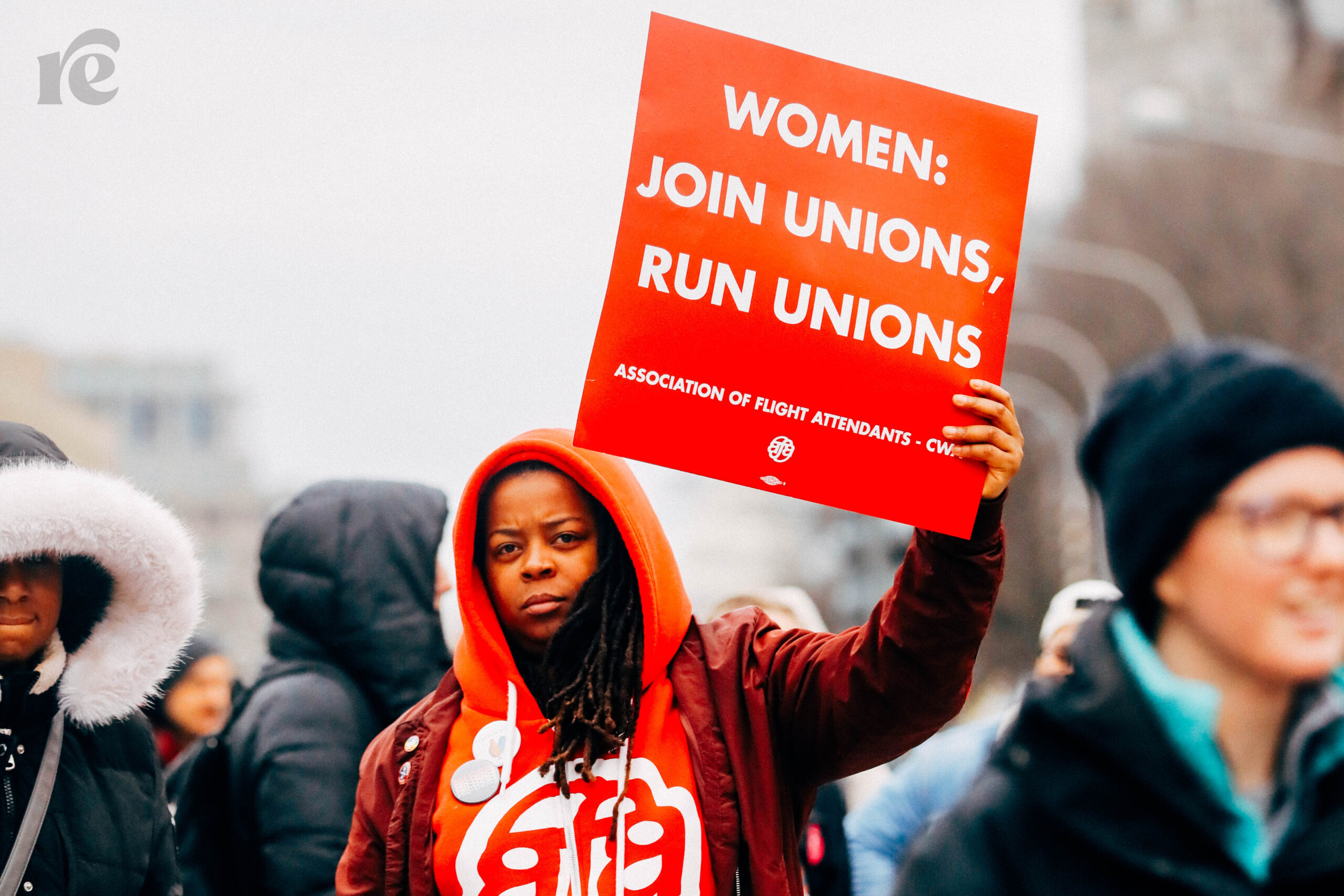 Black woman in red sweatshirt holds red sign that reads Women: Join Unions, Run Unions