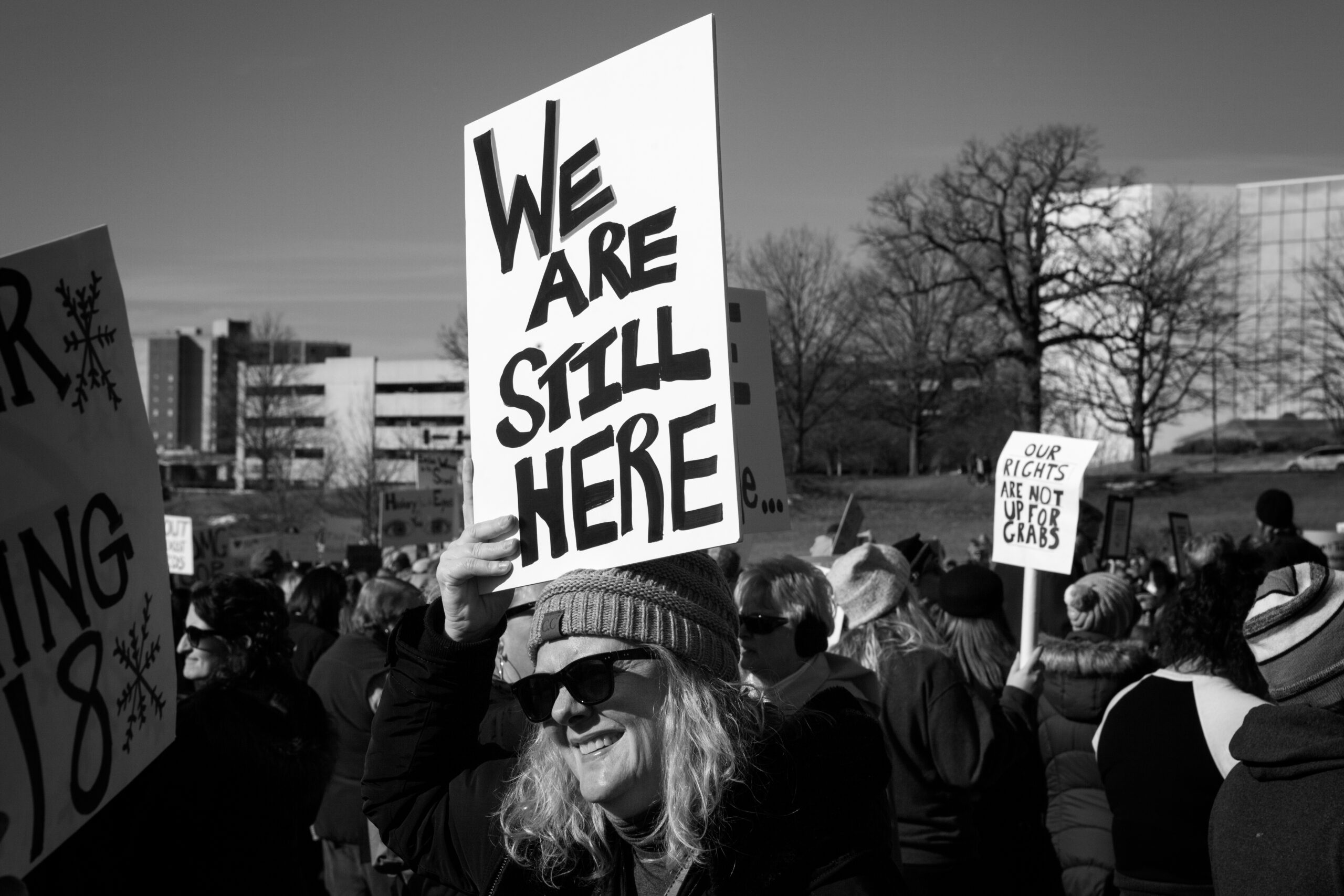 Black and white photo of a white woman holding a sign that reads We are still here
