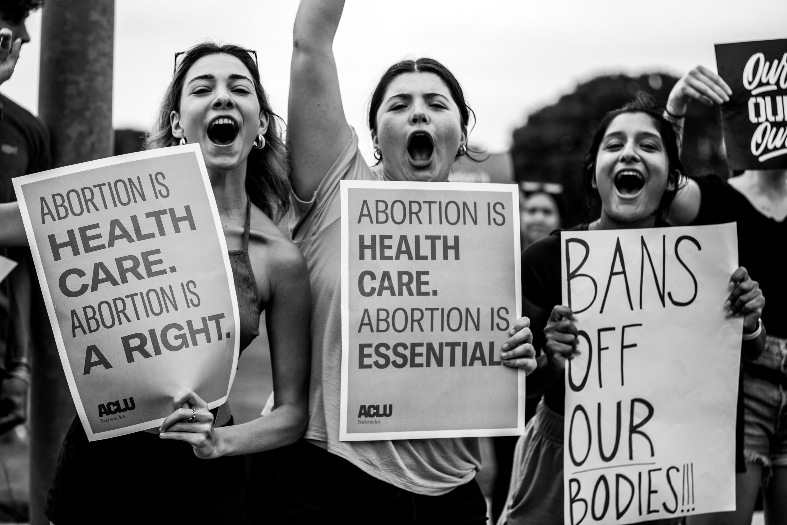 Three protesters hold up signs that read (from left to right): "Abortion is health care. Abortion is a right.", "Abortion is health care. Abortion is essential."And "Prohibitions of our body!!!"