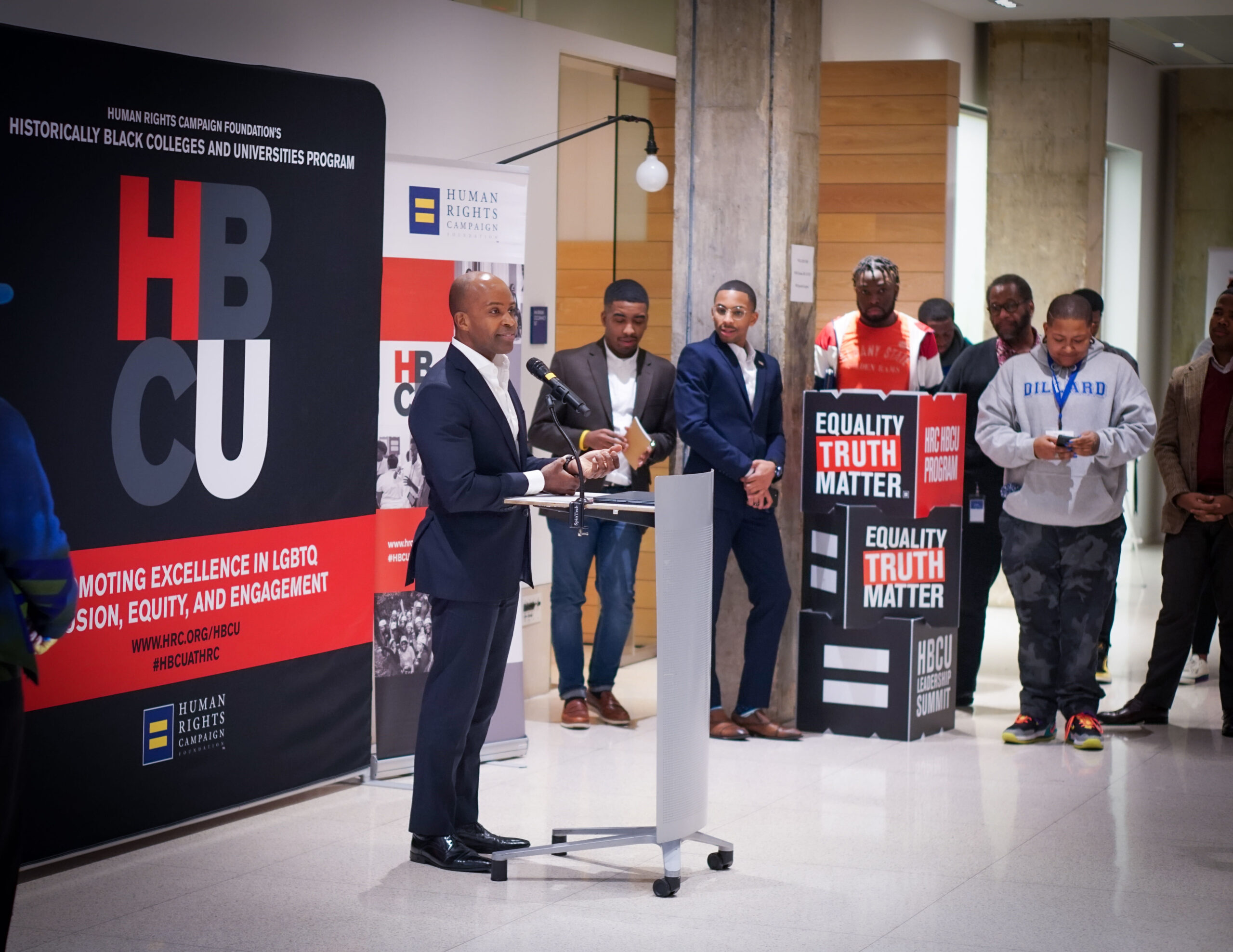 A man wearing a suit speaks at a podium at the Human Rights Campaign HBCU Leadership Summit Opening Reception. A handful of men stand a few feet away watching.