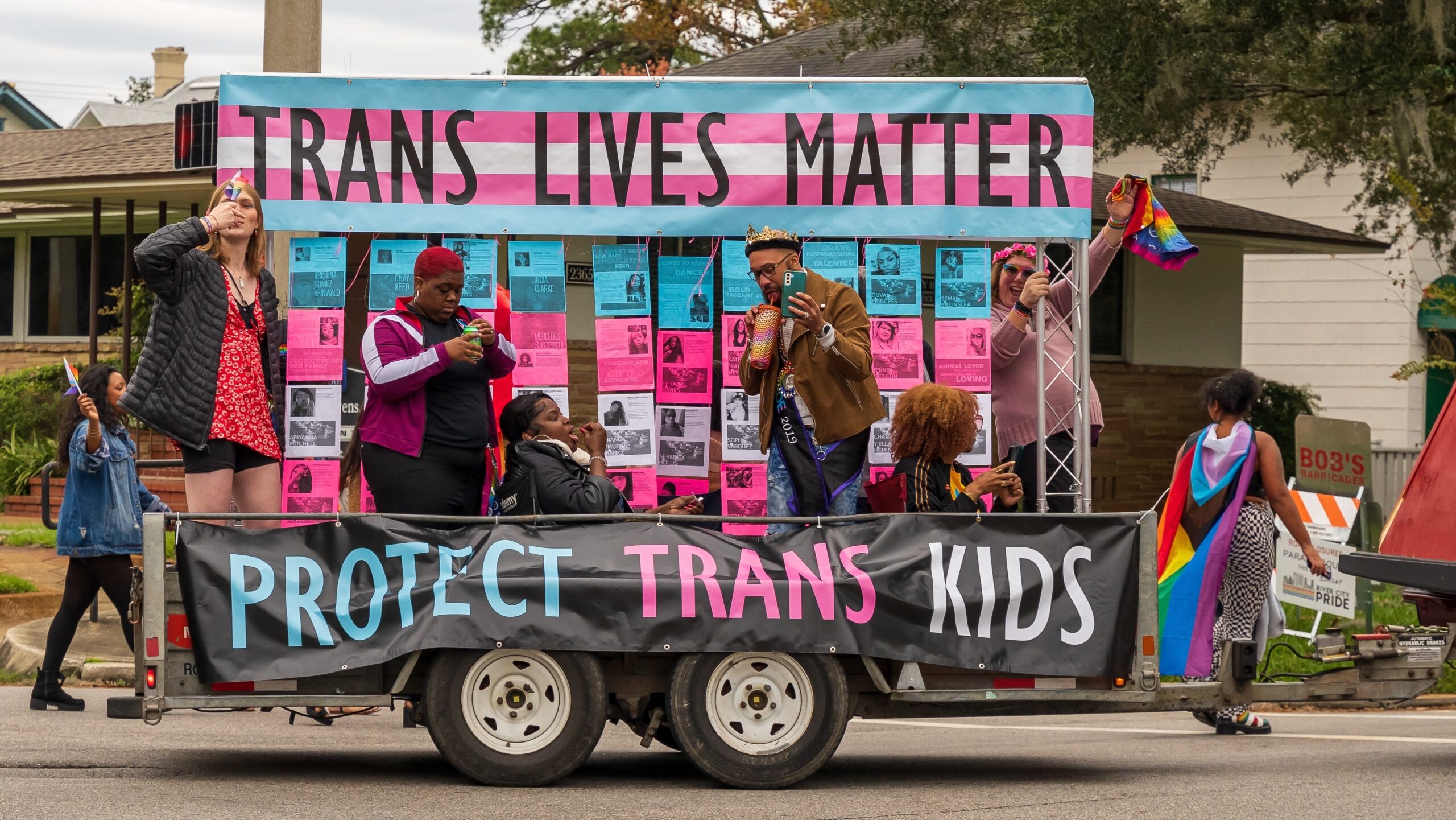 Six people on a parade float with colors from the trans pride flag and banners reading "trans lives matter" and "protect trans kids."