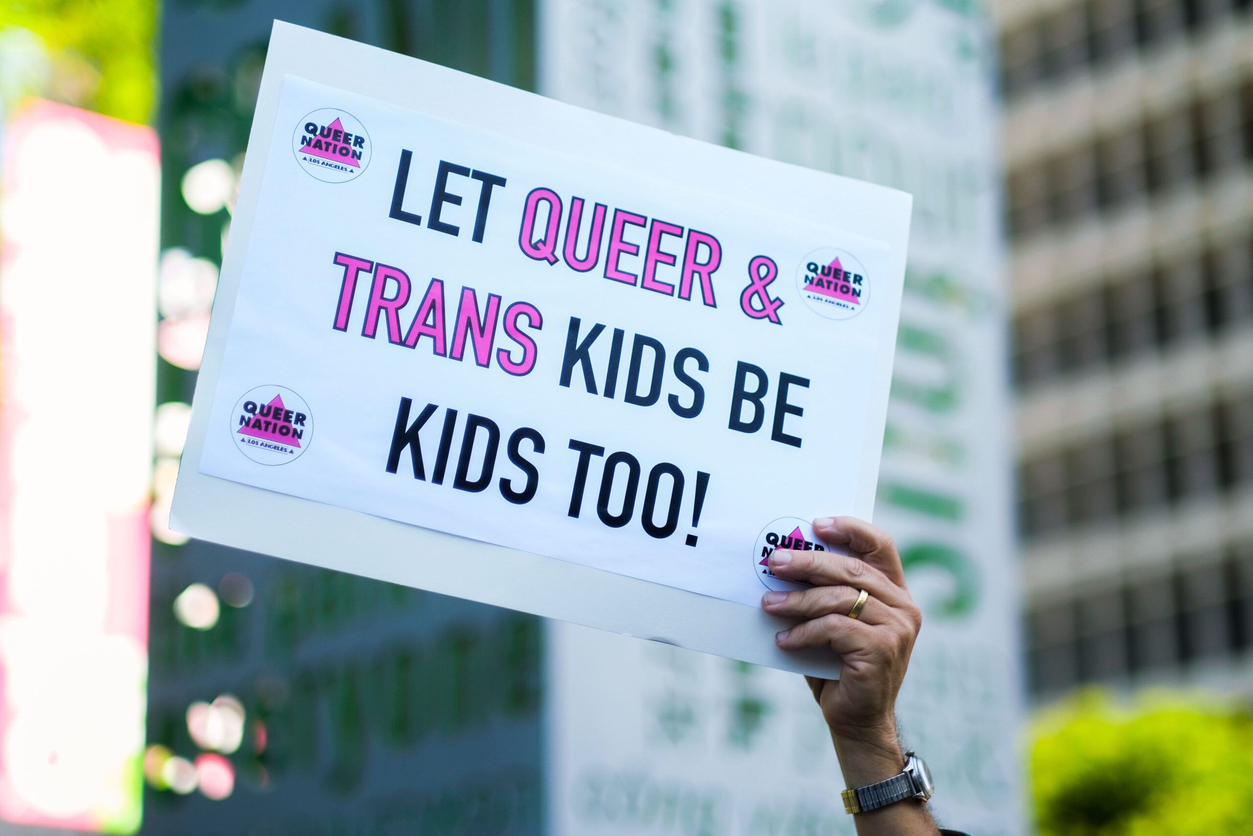 A hand holding a sign that reads "let queer & trans kids be kids too!"